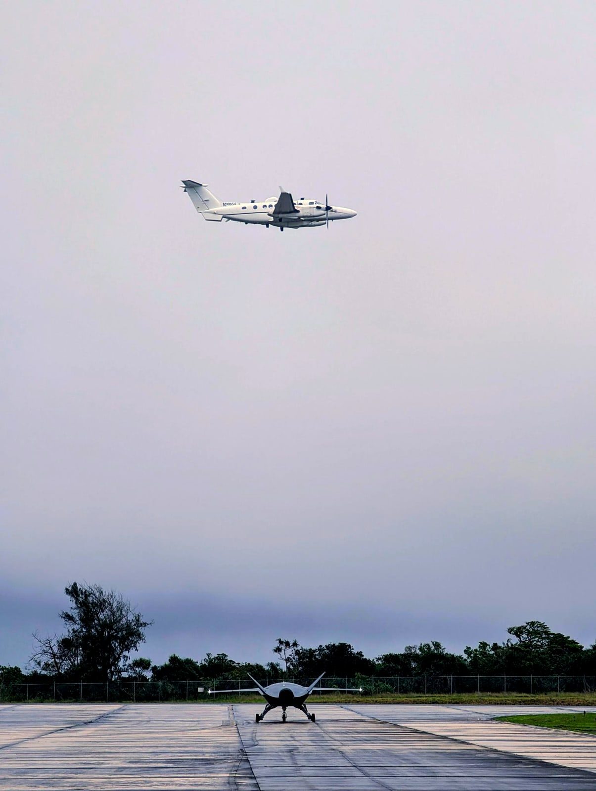 King Air 350ER in a low pass over an uncrewed aircraft parked on a wet island taxiway during a joint exercise